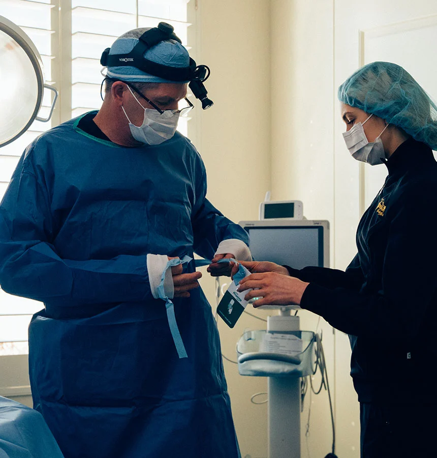 Two medical professionals in blue surgical attire are in a brightly lit medical room. Dr. Urban, a male surgeon, is adjusting the straps of his gown while a female colleague assists him. Both are wearing surgical masks and head coverings. A medical device with a screen is visible behind them, along with large windows with white blinds and a partially visible surgical light fixture. - Facial Trauma in West Jordan, UT