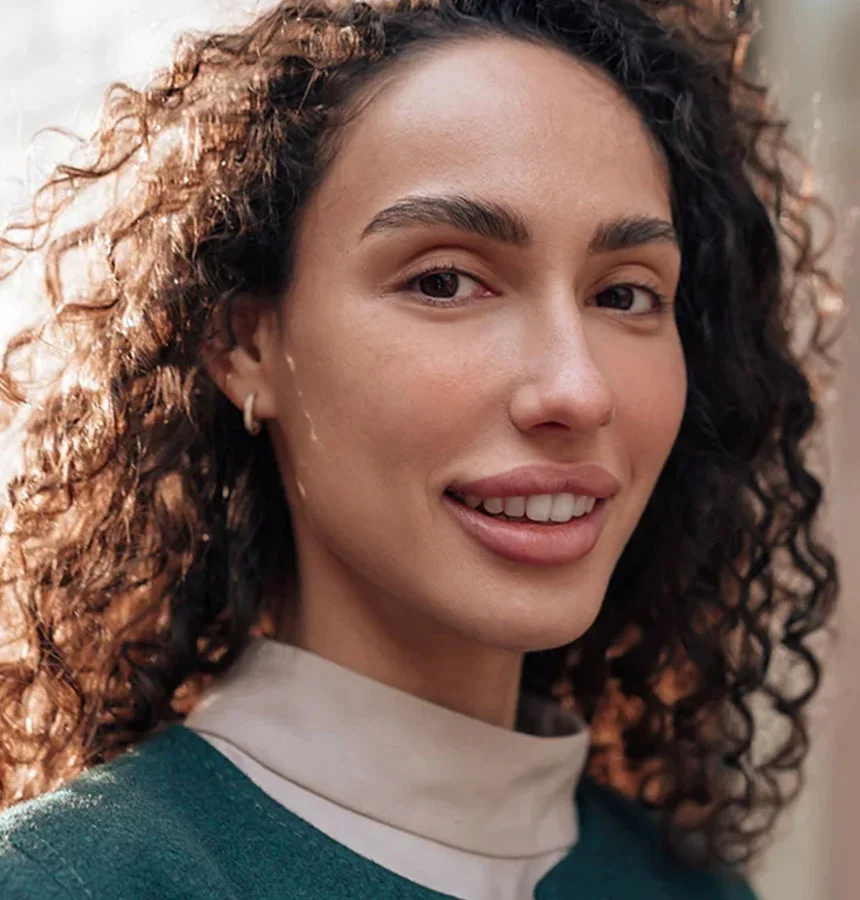 A light-skinned woman with curly, light brown/auburn hair smiles brightly in this close-up. She wears small hoop earrings and a teal or forest-green top layered over a cream-colored collared shirt. Her teeth are visible, and her eyebrows are well-defined. The background is softly blurred. - Impacted Canines in West Jordan, UT