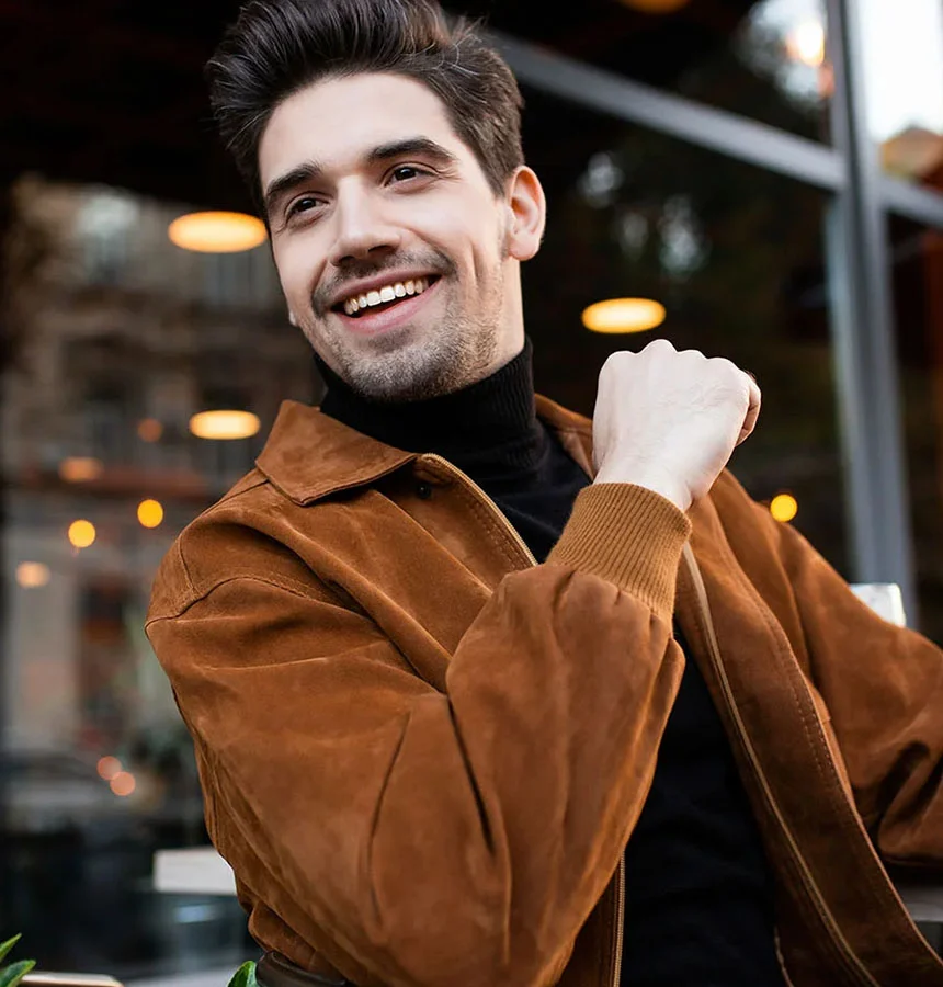 A man with short dark hair smiles as he looks off to his right. He wears a light brown suede jacket over a black turtleneck, with one hand resting on the other. The background is a softly blurred outdoor café with warm lighting and a dark-paneled window. The photo is taken at eye level. - Jaw Surgery in West Jordan, UT