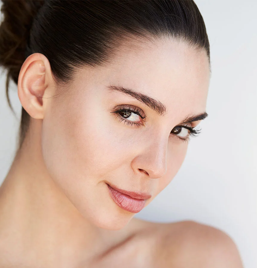 Close-up of a woman with dark brown hair pulled back in a bun, revealing her smooth, light-toned skin. She gazes slightly off-center with a calm, attentive expression. Her well-defined eyebrows, light pink lips, and softly parted smile highlight her natural features. The plain white background keeps the focus on her face and upper chest. - Latisse Eyelash Lengthening in West Jordan, UT
