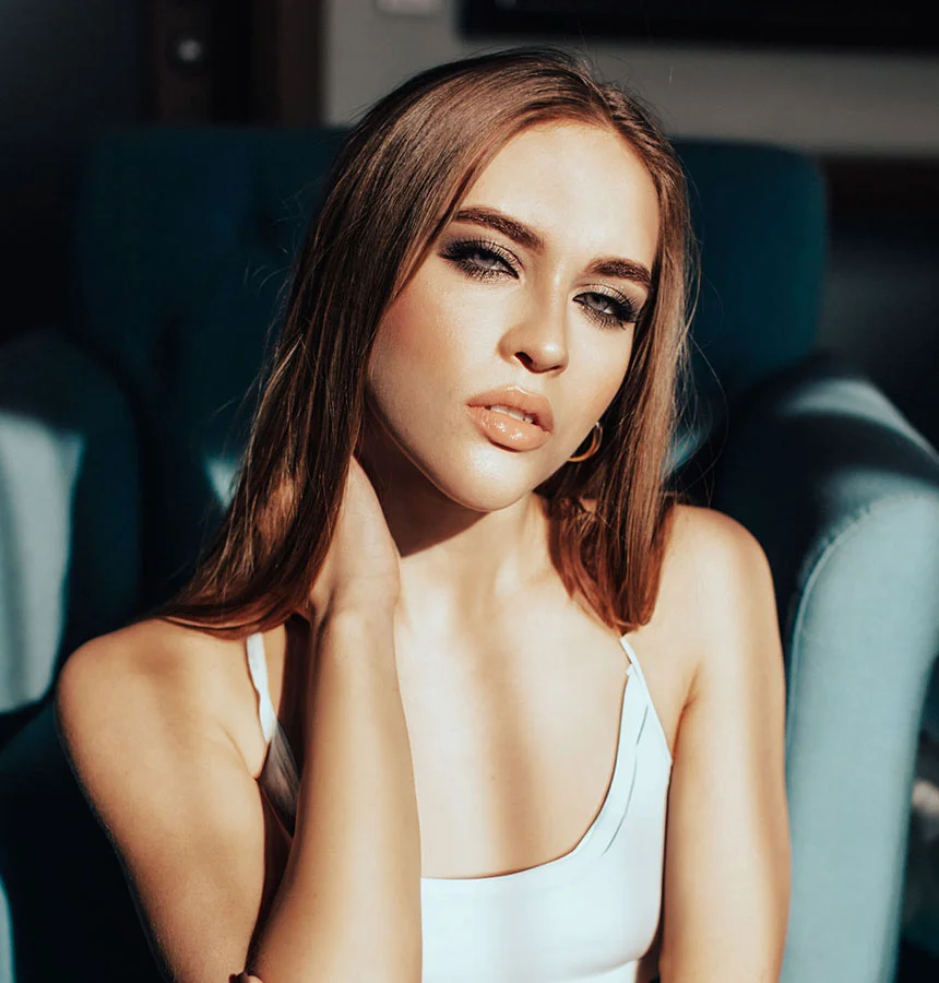 Woman with long straight hair and full lips posing in sunlight while seated on a teal armchair, wearing a white top and subtle makeup with a glossy nude lip. - Lip Fillers in West Jordan, UT