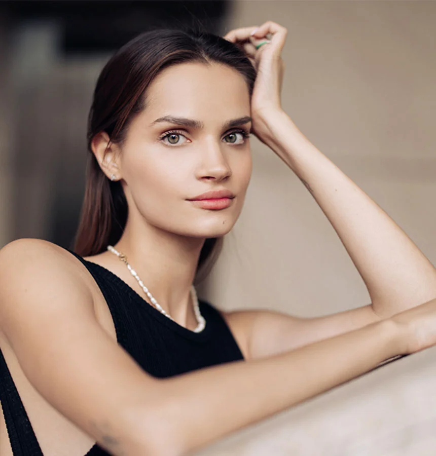 Woman with sleek dark hair and natural makeup resting her head on her hand, wearing a sleeveless black top and a pearl necklace, gazing softly at the camera in a neutral indoor setting. - Lip Lift in West Jordan, UT
