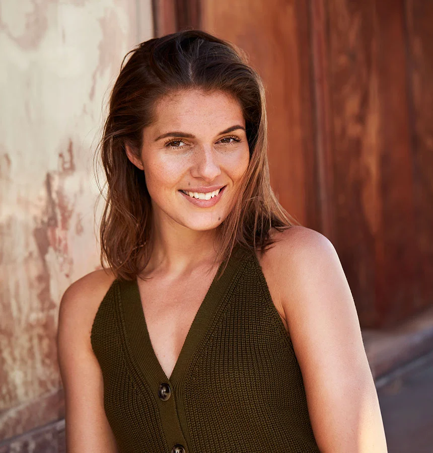 Young woman with shoulder-length brown hair and light freckles smiles at the camera, wearing a deep olive green sleeveless v-neck top. She stands in front of a weathered beige wall with reddish-brown wood in the background, lit by natural light. - Mole Removal in West Jordan, UT