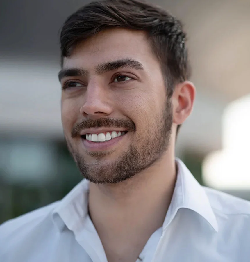 Close-up of a smiling man with short dark brown hair and light stubble, looking upward and to the right. He wears a pale gray collared shirt. The background is softly blurred in light gray-tan tones. - Oral Pathology in West Jordan, UT