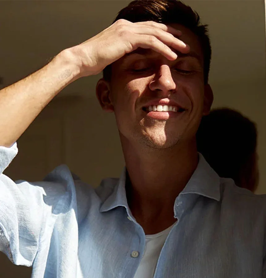 Close-up of a light-skinned man smiling with his eyes closed, shading his forehead with one hand. He wears a light blue linen button-down shirt. Natural light casts soft shadows, highlighting his smile and the shirt’s texture. - Palete Rich Plasma in West Jordan, UT