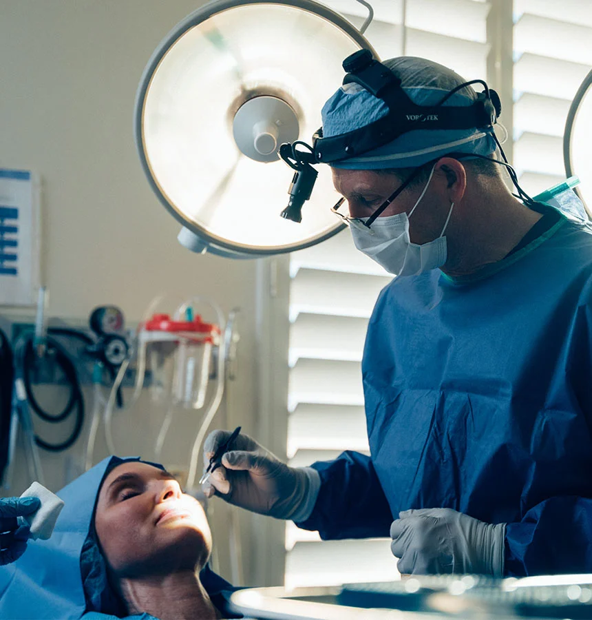 A surgeon in blue surgical gown, mask, and protective eyewear performs a procedure on a patient lying on a table. The surgeon focuses on the patient’s nose and mouth area, holding surgical instruments. Surgical lights and medical equipment are visible in the background. The patient wears a light blue surgical cap. - Pre Prosthetic Surgery in West Jordan, UT