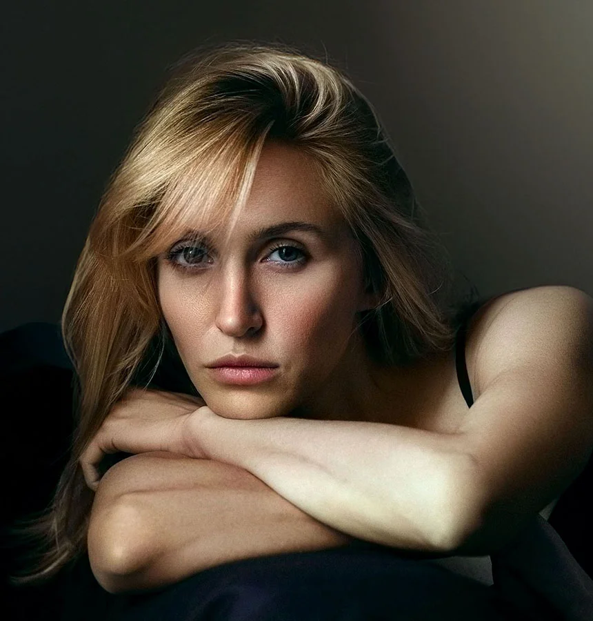 A woman with light blonde hair lies on a bed, resting her head on her arms with legs slightly extended. She has a relaxed, pleasant expression. White bed linens and soft natural light highlight her upper body and face. The background shows slightly blurred window curtains. - RF Microneedling in West Jordan, UT