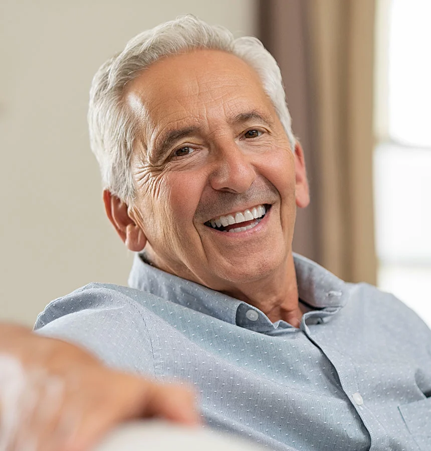 Close-up of a light-skinned senior man with short, graying hair and a broad, toothy smile. His face shows natural wrinkles and he wears a light gray, button-down shirt. The background is softly blurred in beige and cream tones, keeping focus on his face and upper torso. - Stem Cell in West Jordan, UT