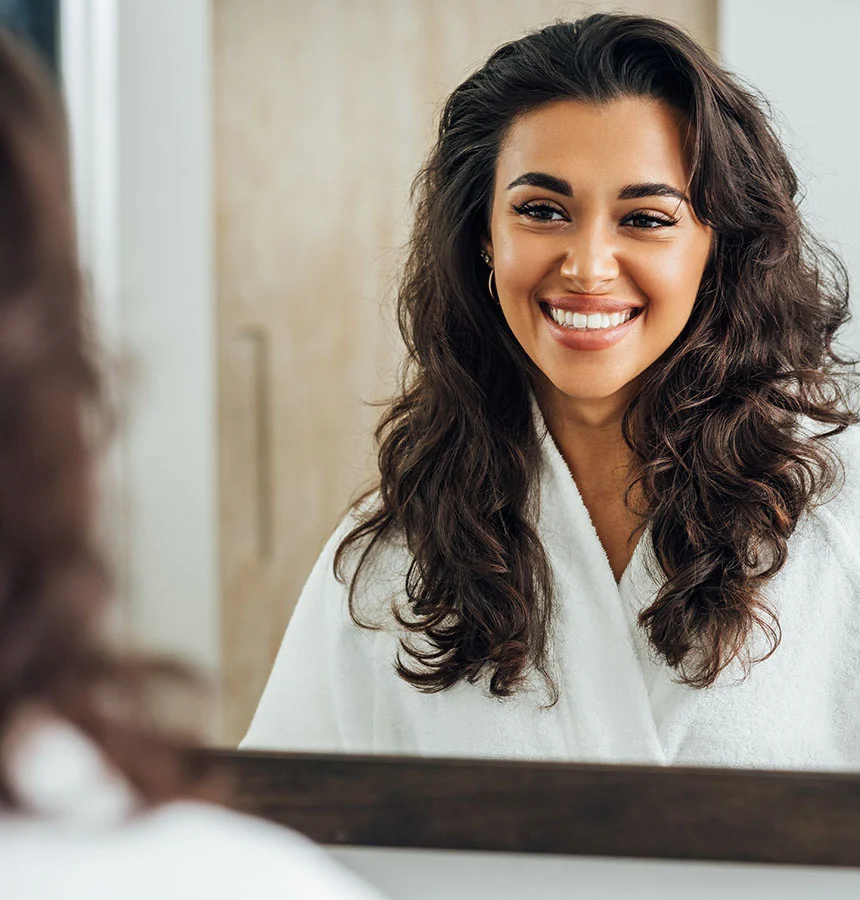 Woman with long wavy dark hair smiling at her reflection in a mirror, wearing a white robe in a bright bathroom setting. - Wisdom Teeth in West Jordan, UT